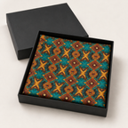 A square black gift box with its lid off, displaying a folded fabric or paper product with a colorful geometric pattern in teal, brown, and gold on a light neutral background.