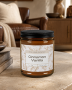 A jar candle labeled 'Cinnamon Vanilla' by Real Cool Company sits on a stone table in a warm, rustic living room setting with a leather sofa and wooden furniture in the background.