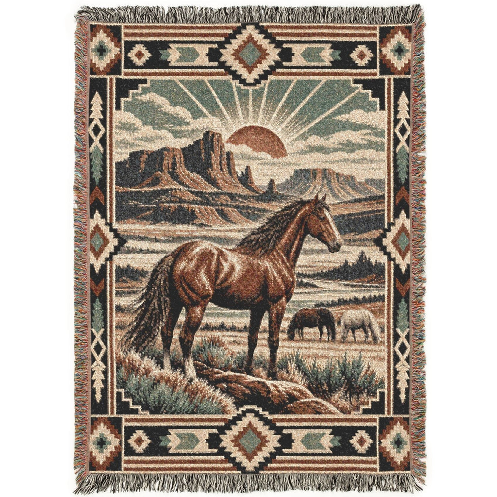 A rectangular woven throw blanket with fringe on all sides, featuring a western landscape scene with a brown horse in the foreground, a smaller animal in the distance, desert mountains, and a sunburst. The border includes geometric patterns in beige, brown, black, and muted green.