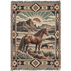 A rectangular woven throw blanket with fringe on all sides, featuring a western landscape scene with a brown horse in the foreground, a smaller animal in the distance, desert mountains, and a sunburst. The border includes geometric patterns in beige, brown, black, and muted green.