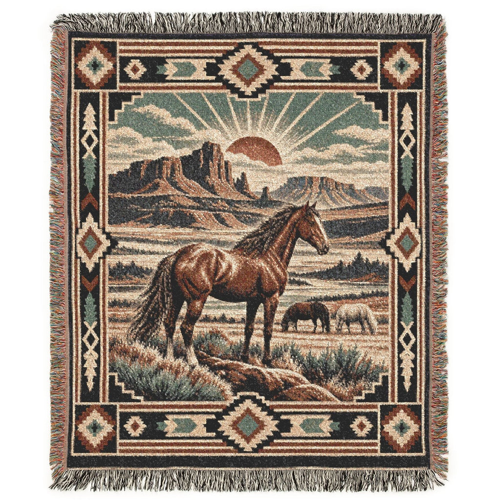 A woven throw blanket with fringe, featuring a western landscape scene with a brown horse in the foreground, another horse in the background, desert mountains, and a sunburst motif, surrounded by a Southwestern geometric border.
