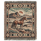 A woven throw blanket with fringe, featuring a western landscape scene with a brown horse in the foreground, another horse in the background, desert mountains, and a sunburst motif, surrounded by a Southwestern geometric border.