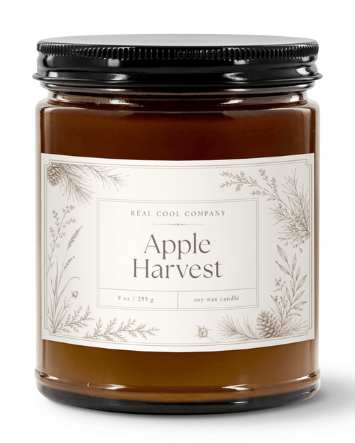 A 9 oz soy wax candle in a amber glass jar, a cream label with botanical illustrations, and a black metal lid. The label reads 'REAL COOL COMPANY' and 'Apple Harvest'.