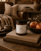 An amber glass jar candle with a black lid and a cream label reading 'Apple Harvest' sits on stacked books on a wooden table in a cozy living room setting with a leather sofa, throw blanket, and apples in a bowl.