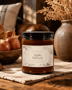 A amber glass jar candle labeled 'Apple Harvest' by Real Gone Company, topped with a black lid, styled on a rustic table with apples, dried leaves, and a ceramic vase in the background.