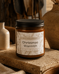 A glass jar candle with soy blend wax and a black lid sits on a stone surface in a warm, rustic setting. The front label reads 'REAL COOL COMPANY Christmas Warmth' with delicate botanical line art.