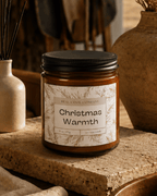 A glass jar candle with soy blend wax and a black lid sits on a stone surface in a warm, rustic setting. The front label reads 'REAL COOL COMPANY Christmas Warmth' with delicate botanical line art.