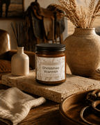 A jar candle labeled 'Christmas Warmth' by Red Cedar Company sits on a stone slab in a rustic, warm-toned interior setting with dried grasses, a ceramic vase, and wooden decor.