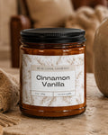A 9 oz soy candle in an amber glass jar with a black lid and a cream label reading 'REAL COOL COMPANY Cinnamon Vanilla', styled on a stone surface in a cozy living room setting with a fireplace, knitted blanket, and vase in the background.