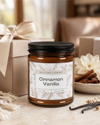 A jar candle labeled 'Cinnamon Vanilla' by Real Cool Company, in an amber glass jar with a black lid, styled on a tabletop with a kraft gift box, vanilla beans, and cinnamon sticks.