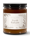 A 9 oz soy wax candle in a clear glass jar, a black metal lid, and a cream label featuring pine illustrations. The label reads 'REAL COOL COMPANY Fresh Balsam'.