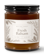 A 9 oz soy wax candle in a clear glass jar, a black metal lid, and a cream label featuring pine illustrations. The label reads 'REAL COOL COMPANY Fresh Balsam'.