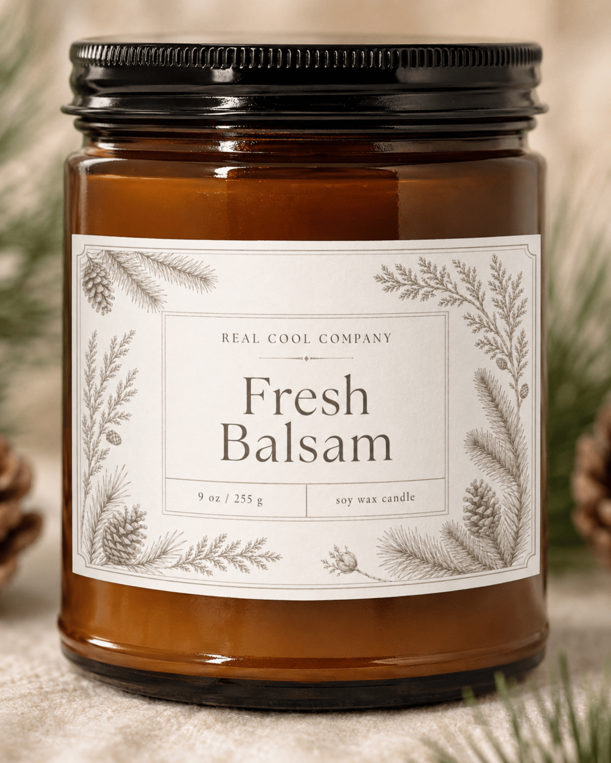 A 9 oz soy wax candle in an amber glass jar with a black metal lid and a white botanical label reading 'Fresh Balsam' by the Real Cool Company, styled with pinecones and evergreen branches.