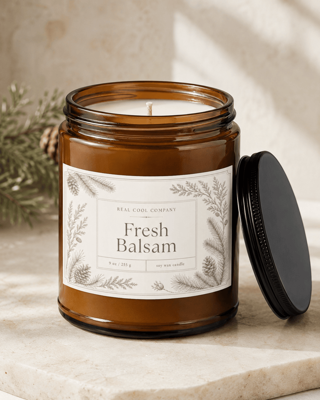 An amber glass jar candle labeled Fresh Balsam by Real Cool Company, shown with its black lid resting beside it on a light stone surface.