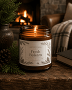 A lit amber glass jar candle labeled 'Fresh Balsam' by Real Cool Company sits on a rustic wooden surface in a cozy indoor setting with a fireplace, evergreen branches, and a plaid blanket in the background.
