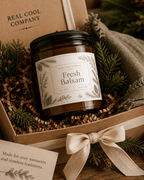 A jar candle labeled 'Fresh Balsam' by Real Cool Company, presented in a kraft gift box with pine branches, a ribbon bow, and filler paper.