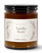 A jar candle labeled 'Vanilla Bean' by Real Cool Company, featuring vanilla bean wax in a clear glass jar with a black metal lid and a cream-colored botanical label.