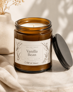 An amber glass jar candle labeled 'Vanilla Bean' by Real Cool Co. Company, shown open with its black lid resting beside it on a light stone surface.