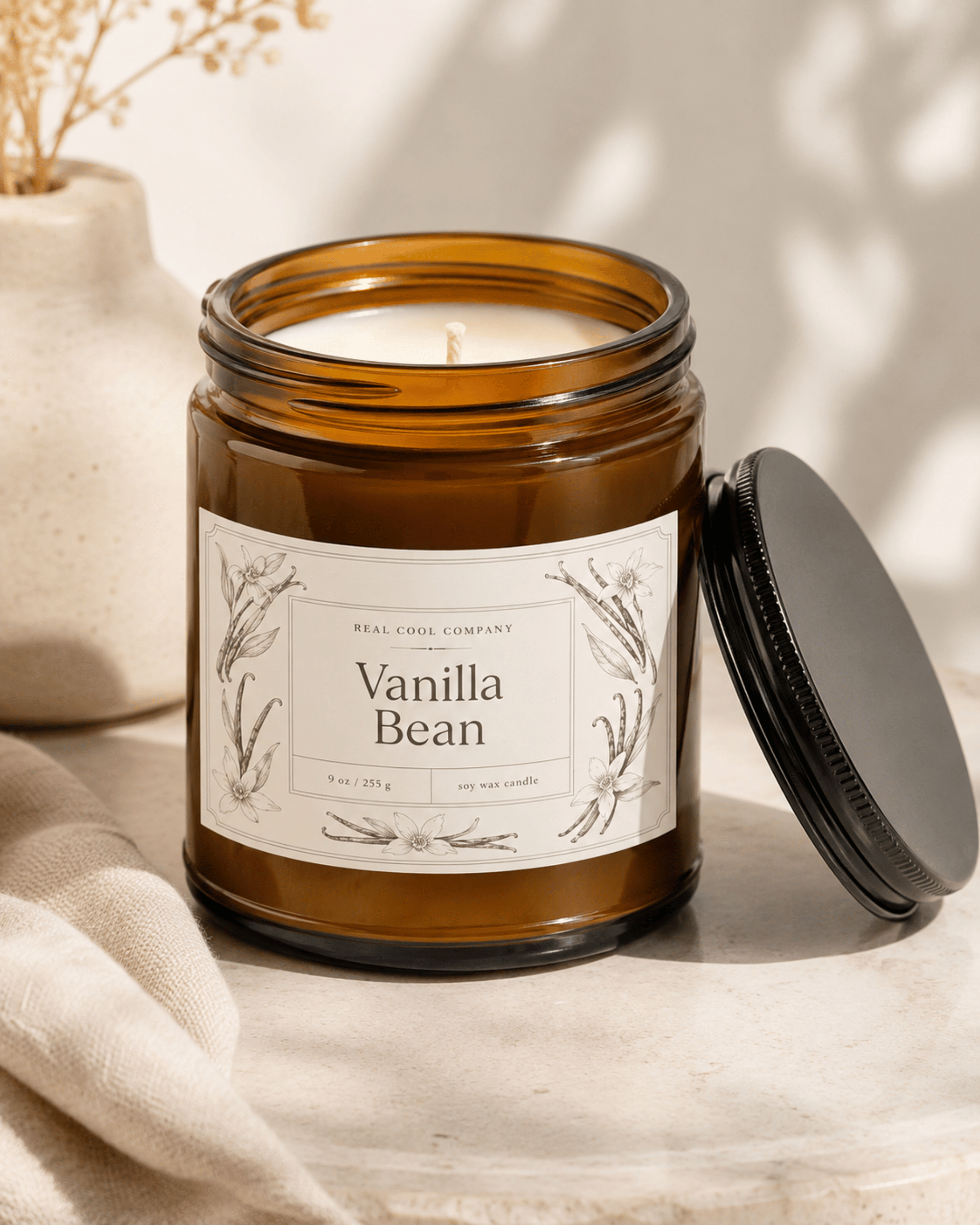 An amber glass jar candle labeled 'Vanilla Bean' by Real Cool Co. Company, shown open with its black lid resting beside it on a light stone surface.