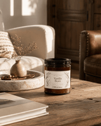 A jar candle labeled 'Vanilla Bean' sits on a rustic wooden table in a cozy living room setting with a cream sofa, leather armchair, and dried flowers in the background.