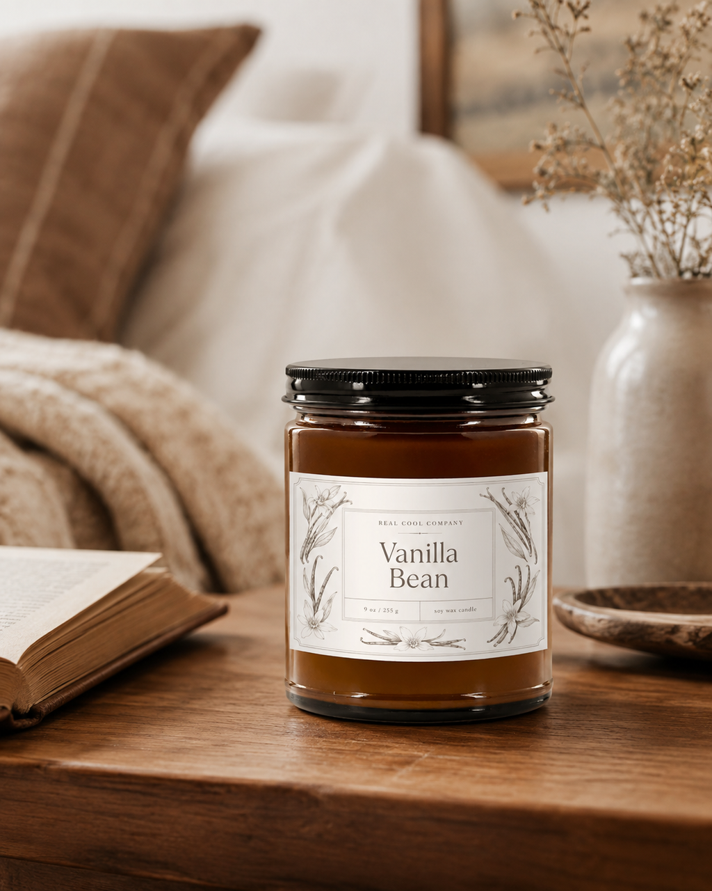 A glass jar candle labeled 'Vanilla Bean' by Real Good Candle Co. sits on a wooden table in a cozy bedroom setting with a blanket, book, and vase in the background.