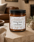 A 9oz amber glass jar candle with a black metal lid and a white paper label reading 'White Sage & Lavender' by Real Cool Company, styled on a stone surface in a cozy living room setting.