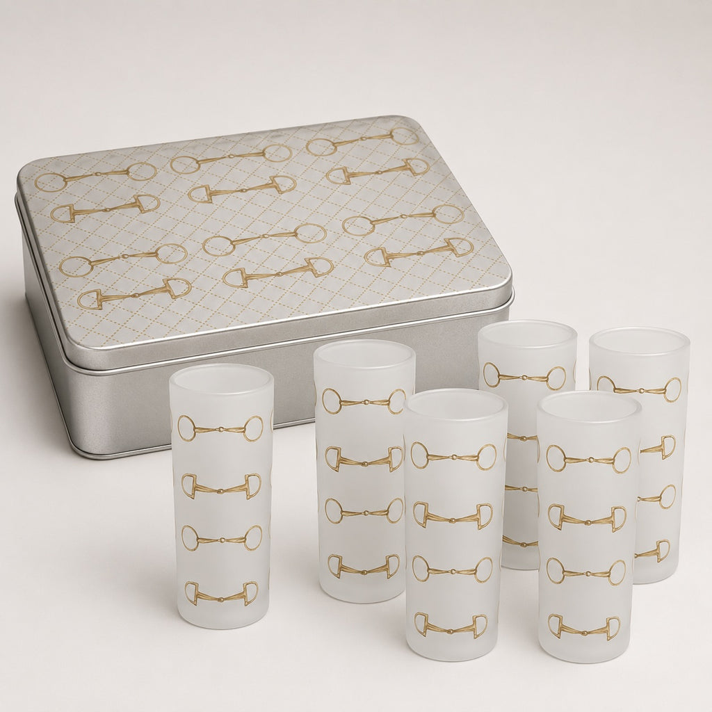 Six frosted glass shot glasses with a gold horsebit pattern, displayed in front of a silver metal tin with a matching horsebit design on a light beige background.