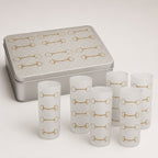 Six frosted glass shot glasses with a gold horsebit pattern, displayed in front of a silver metal tin with a matching horsebit design on a light beige background.