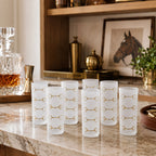 Six frosted glass shot glasses with gold horse-bit patterns arranged on a marble bar counter in a warm, stylish bar setting.