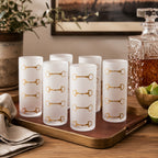 Six frosted glass tumblers with gold horsebit motifs, arranged on a brown tray in a styled bar setting with a decanter, lime, and greenery.