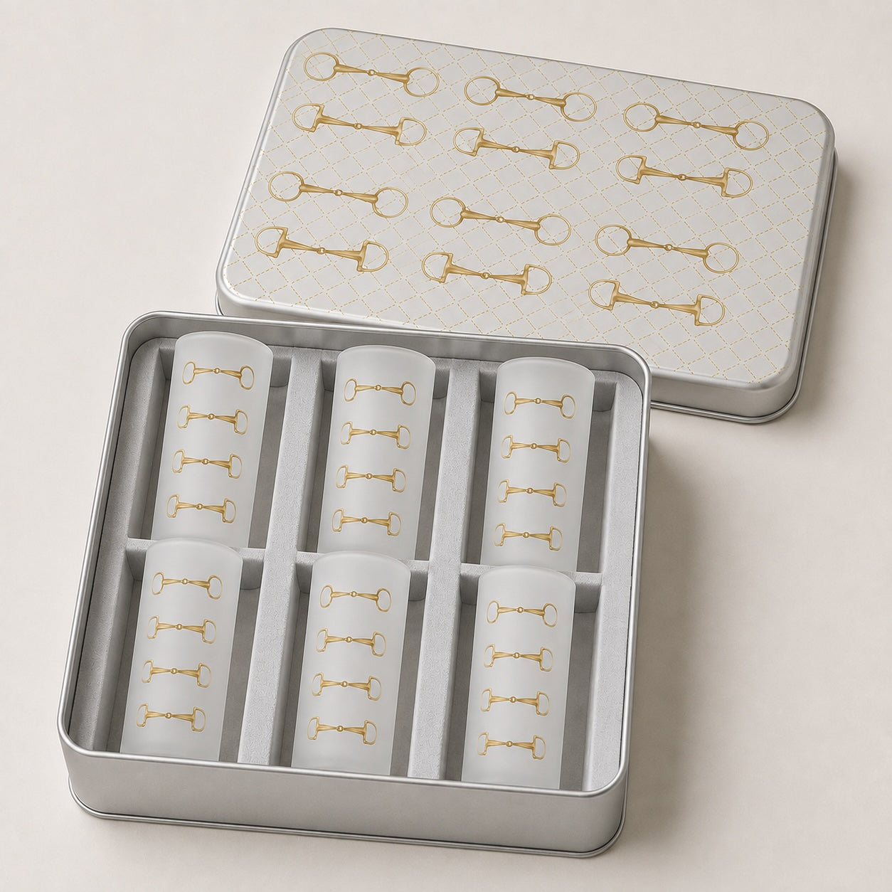 A silver metal gift tin with its lid placed behind it, containing six frosted glass shot glasses. The lid and tubes are decorated with a repeating gold horsebit pattern on a light background.