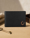 A black textured leather bifold wallet with a small brown horseshoe logo on the bottom right, displayed on a light wood tabletop with a brass key, folded beige cloth, and a brown leather belt in the background.