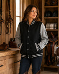 A woman stands in a rustic indoor setting wearing a varsity-style jacket with a black body and gray sleeves. The jacket has white striped ribbed trim, white snap buttons, and 'RANCH CLUB' printed vertically on the sleeve.
