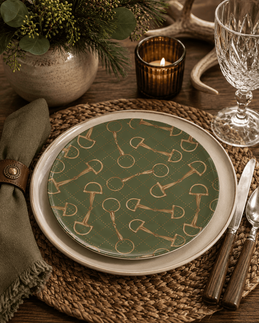 A styled table setting featuring a green china dinner plate with a gold horse-bit pattern, layered over a cream charger plate, on a woven placemat with a green napkin, knife, spoon, and amber candle.