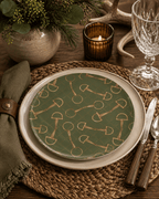 A styled table setting featuring a green china dinner plate with a gold horse-bit pattern, layered over a cream charger plate, on a woven placemat with a green napkin, knife, spoon, and amber candle.