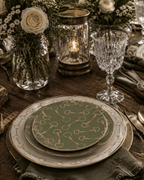 A styled table setting featuring a stack of china plates with a sage green and gold horse-bit pattern, set on a rustic wooden table with a glass goblet, candle holder, and floral arrangements.