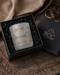 A frosted glass candle with gold horse-bit motifs, presented in a black gift box with a black lid, styled on a rustic wooden table with leather and fabric props.