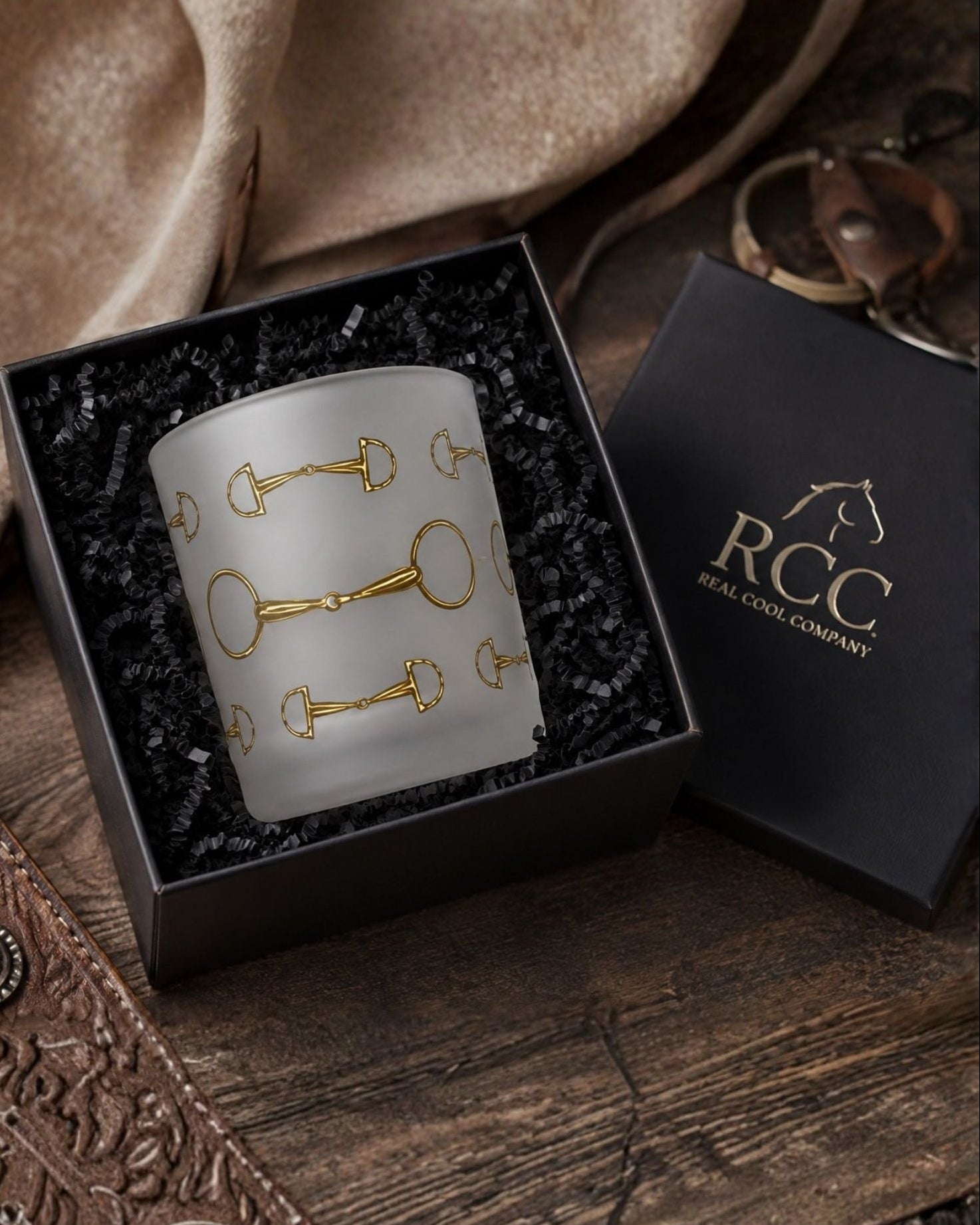 A frosted glass candle with gold horse-bit motifs, presented in a black gift box with a black lid, styled on a rustic wooden table with leather and fabric props.