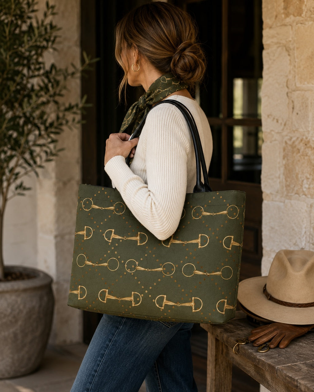A woman in a white ribbed sweater and blue jeans carries an olive green tote bag with a gold horse-bit pattern, standing next to a rustic wooden bench with a beige hat resting on it.