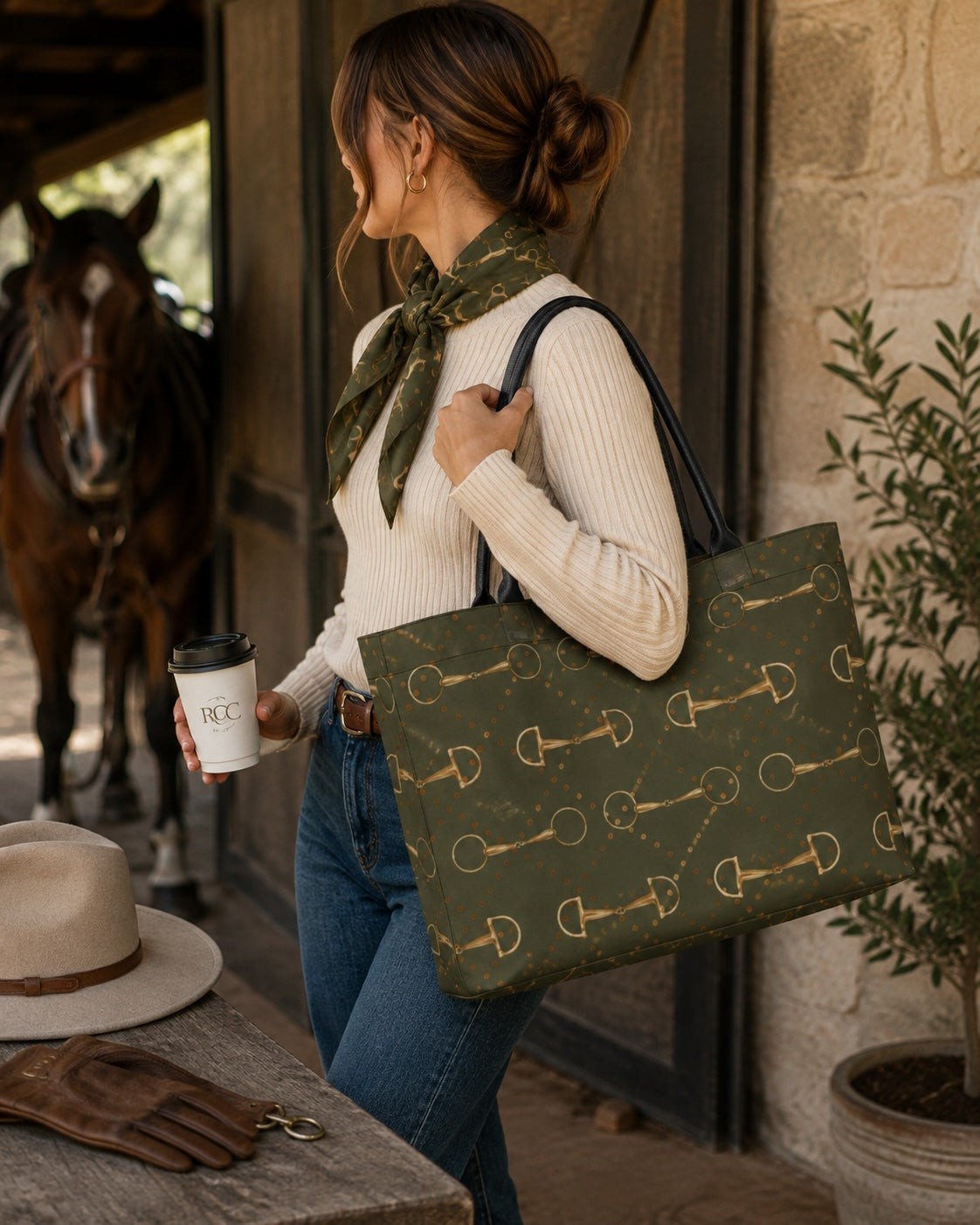 A woman in a stable holds a large olive green tote bag with a gold equestrian pattern. She is wearing a cream ribbed sweater, blue jeans, and a matching olive green neck scarf, with a horse and a rustic stable interior in the background.