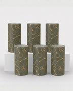 Six cylindrical decorative shot glasses in a muted olive green color, featuring a repeating metallic gold horse bit motif, arranged on a white display riser against a light gray background.