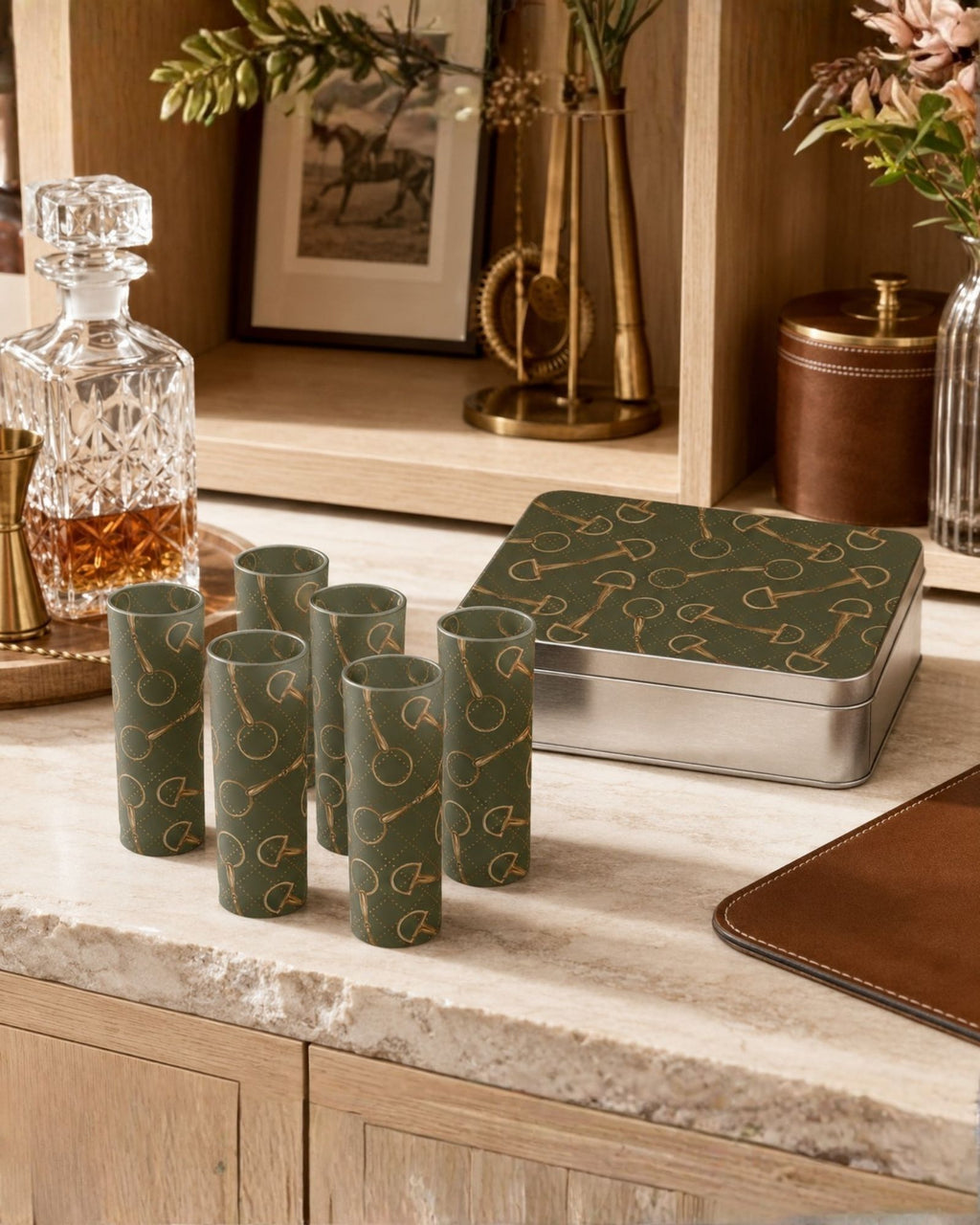 A set of six olive green shot glasses with gold horse-bit patterns, displayed next to a matching metal tin on a marble countertop in a warm, rustic interior setting.