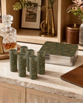 A set of six olive green shot glasses with gold horse-bit patterns, displayed next to a matching metal tin on a marble countertop in a warm, rustic interior setting.
