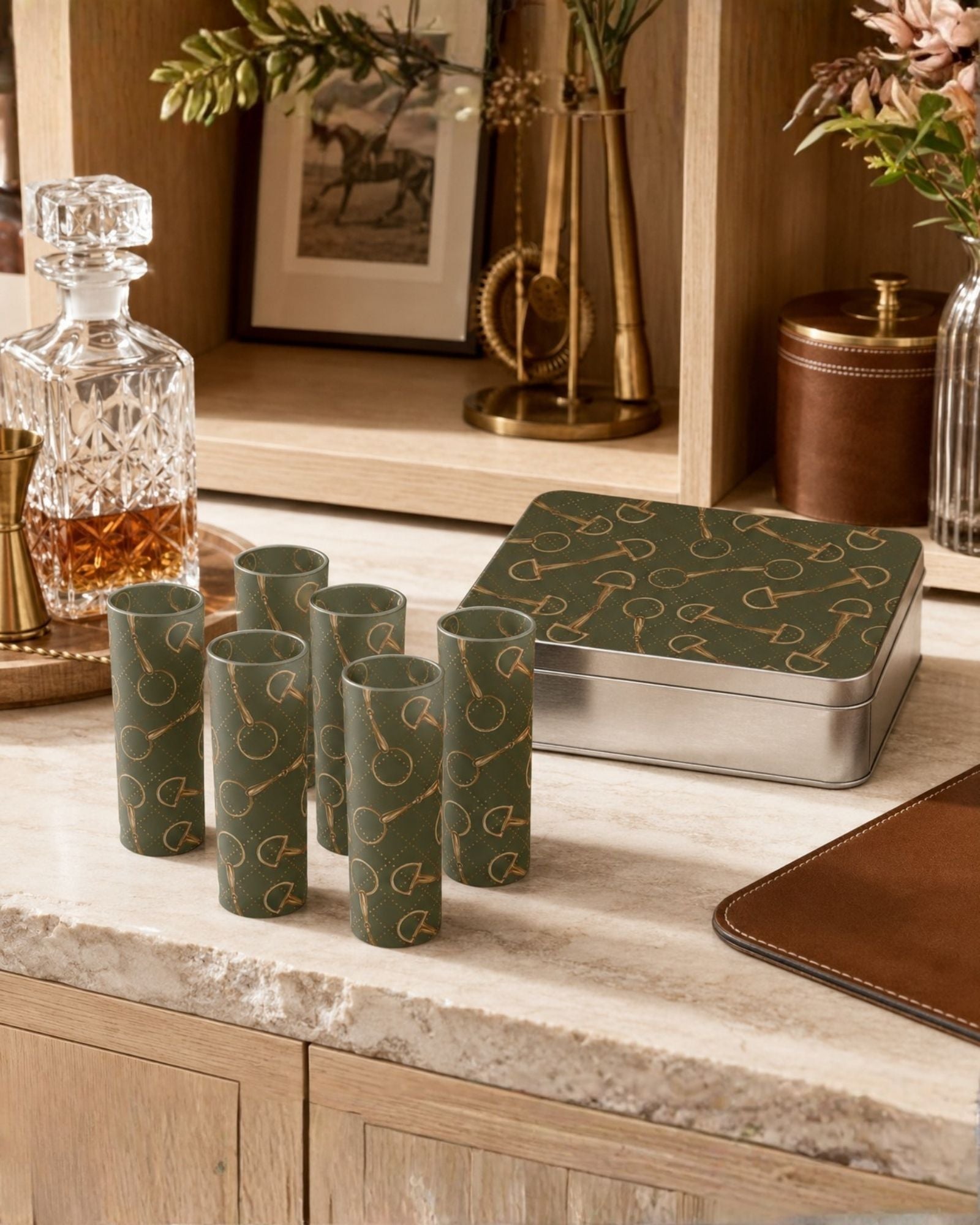 A set of six olive green shot glasses with gold horse-bit patterns, displayed next to a matching metal tin on a marble countertop in a warm, rustic interior setting.