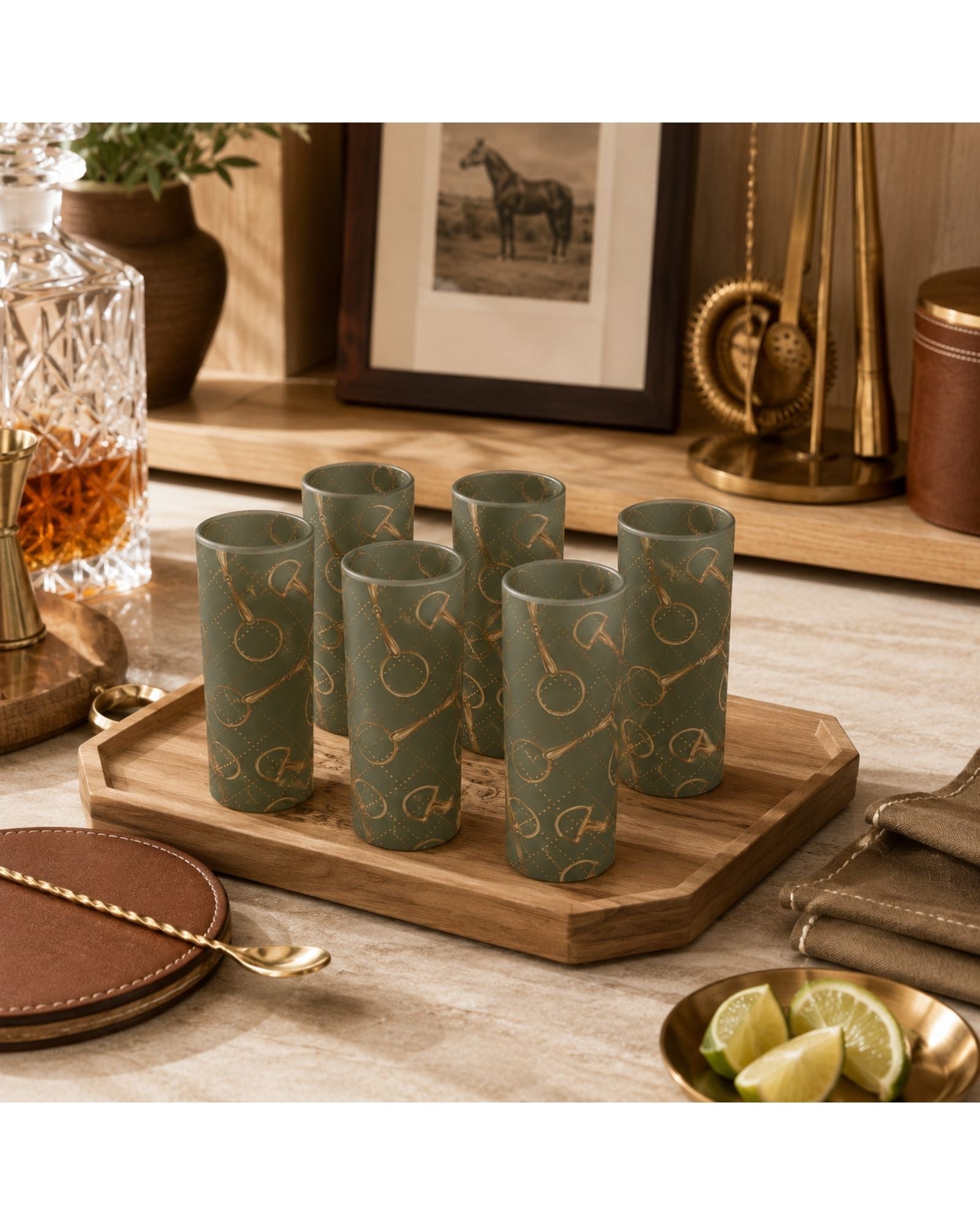 Six olive green gold horse bit pattern shot glasses arranged on a wooden tray, set in a warm, styled ranch home environment with a decanter, framed picture, and lime slices.