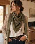 A woman wearing a beige blazer, white shirt, and dark blue jeans models a sage green square scarf with a gold chain-link pattern in a warm, rustic indoor setting.