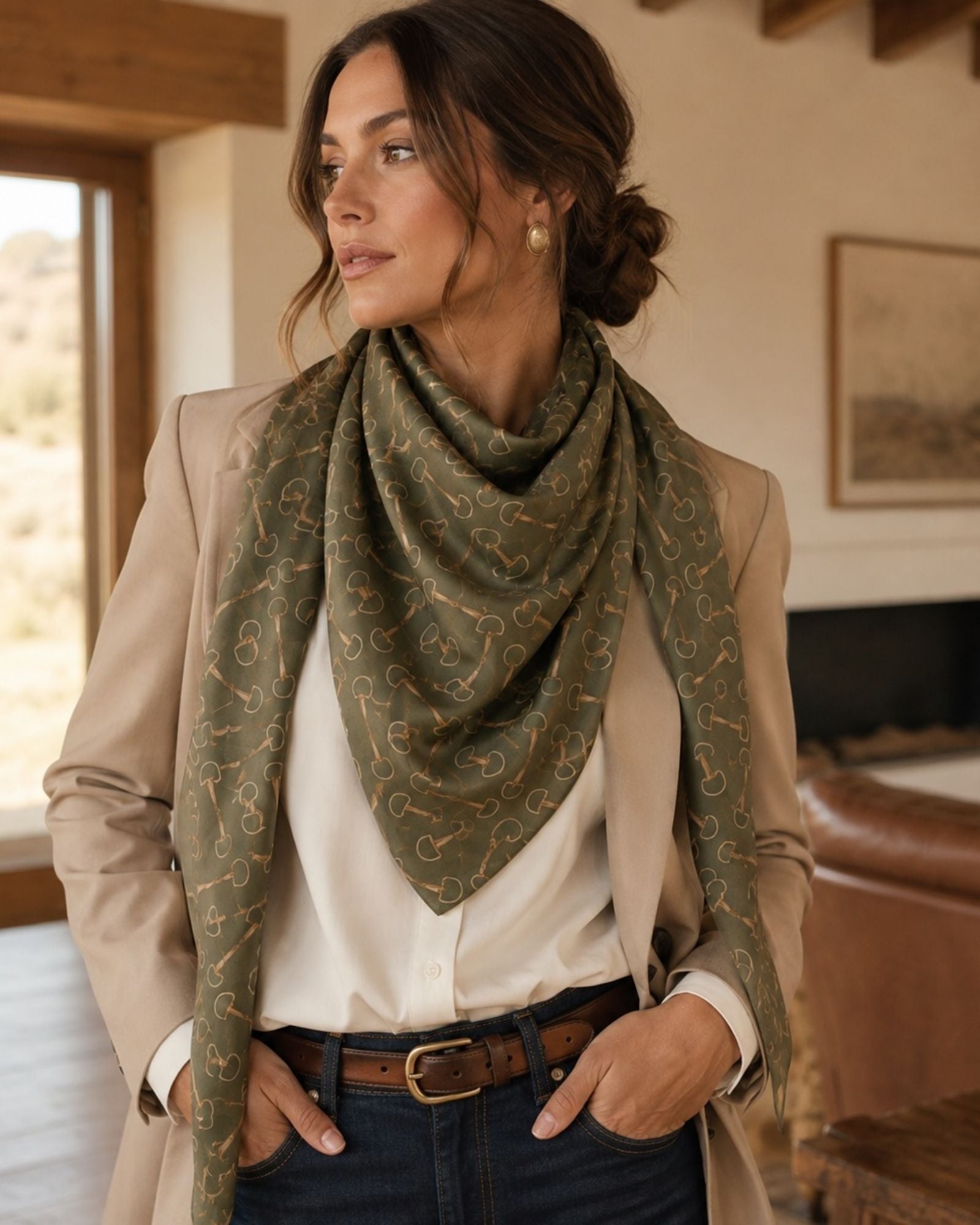 A woman wearing a beige blazer, white shirt, and dark blue jeans models a sage green square scarf with a gold chain-link pattern in a warm, rustic indoor setting.