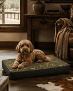 A golden doodle lies on a rectangular dog bed in a rustic living room. The bed has a green and tan horse-and-ribbed patterned top with dark charcoal sides.