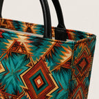 Close-up of a custom designed luxury denim tote bag with black leather handles and a colorful geometric Southwestern-style pattern in teal, rust orange, brown, and cream on a light background.