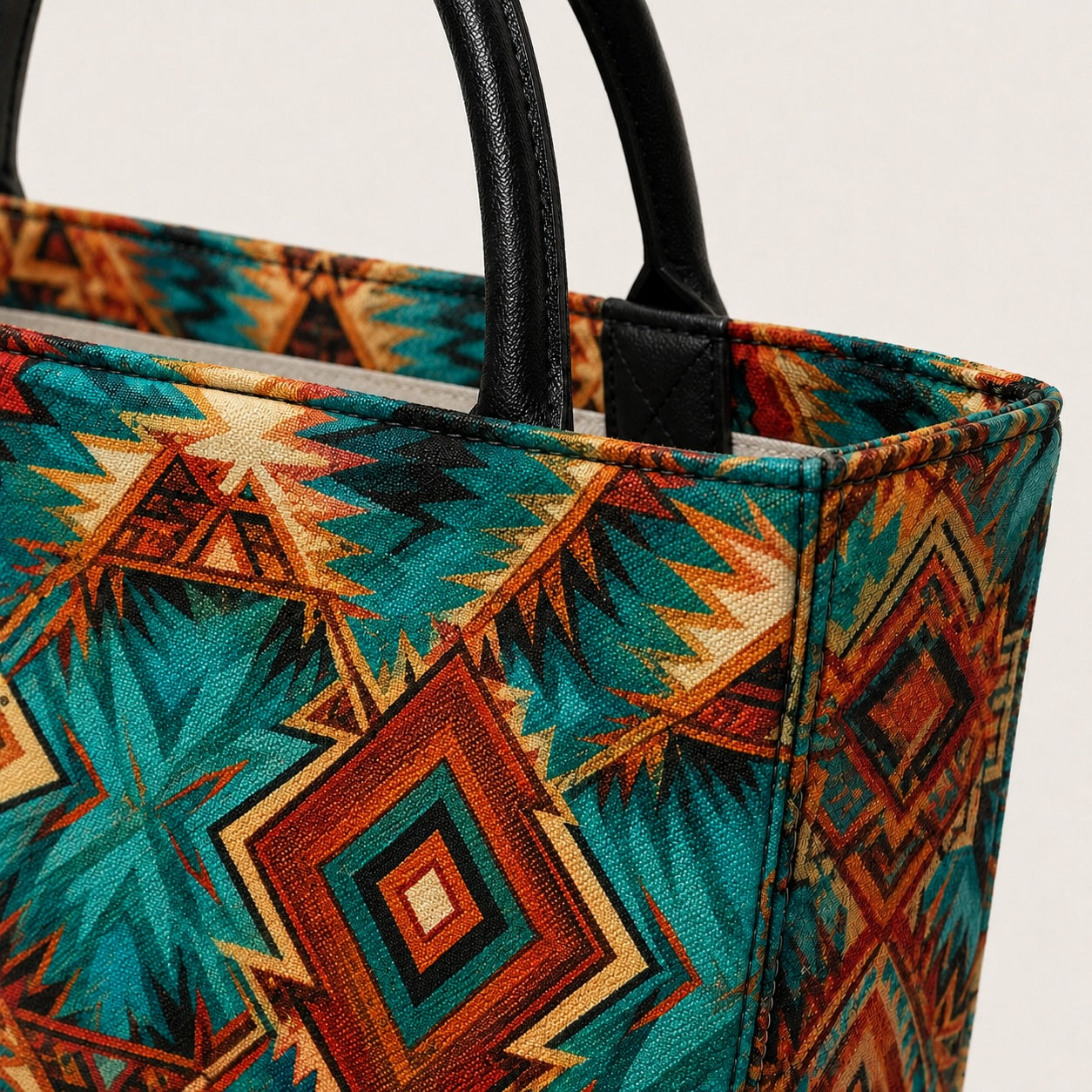 Close-up of a custom designed luxury denim tote bag with black leather handles and a colorful geometric Southwestern-style pattern in teal, rust orange, brown, and cream on a light background.